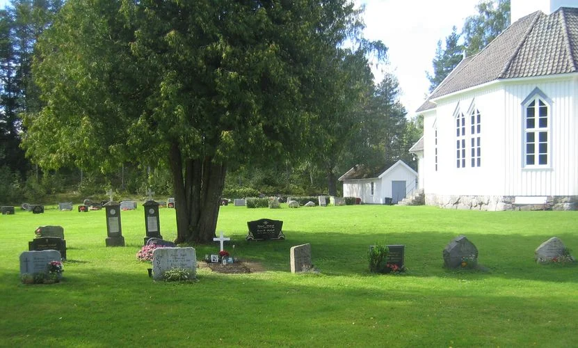 Kilebygda kirke og kirkegård med gravminner. Det er et stort og flott tre mellom gravene. Foto.